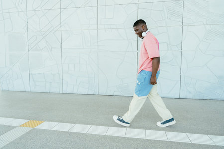 Full height of African man in a pink shirt and blue jeans walks down a sidewalk. He is wearing...