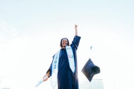 A woman wearing a black graduation gown and a blue cap is holding a diploma. She is smiling and she...