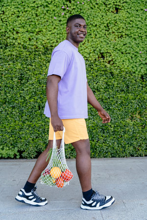 Vertical of African Man Going Grocery Shopping With A Reusable Shopping Bag walks along the green...