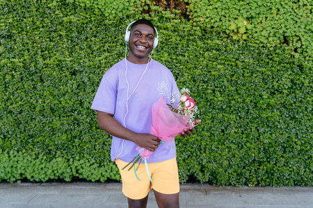 Happy African man with headphones wearing a purple shirt is holding a bouquet of flowers. He is...
