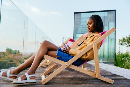 Sad thoughtful African woman is sitting on a wooden chair with her legs crossed on terrace with city view. Phone, social media and communication concept.の写真素材