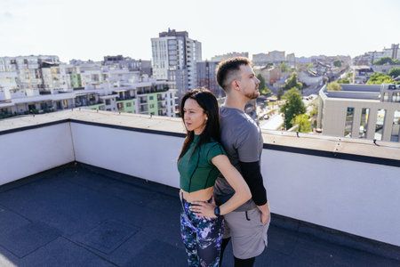 Athletic man and flexible woman standing back to back in morning in rooftop on urban background.