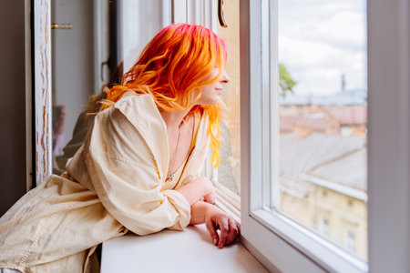 Adult Caucasian woman with colorful red and orange hair looks thoughtfully outside from window, dressed in casual, leans on windowsill, enjoying peaceful moment indoors.の写真素材
