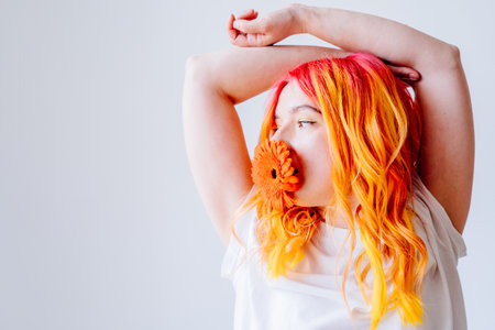 Portrait of young Caucasian woman with bright orange and yellow hair holding orange flower in mouth. Creativity, individuality, vivid colors, unique style. Concepts of self-expression, artistic flair.の写真素材