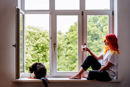 Young woman with colorful hair seated by window holds drink, cat plays nearby. Serene domestic,...