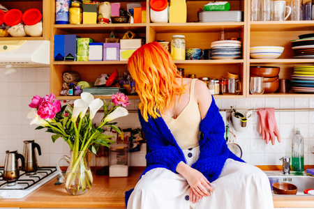 Caucasian woman artist with bright orange hair sits on kitchen counter surrounded by colorful decor and flowers. Creativity, comfort, and joy in everyday life. Personal style, simple pleasures at homeの写真素材