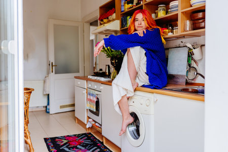 Caucasian young woman with vibrant rainbow hair sitting at counter in cozy kitchen. Female wears blue sweater, domestic lifestyle in artistic and relaxed way. Creativity and everyday life concept.の写真素材