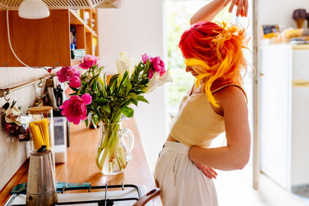 Vibrant young woman with colorful hair dances joyfully in bright kitchen with flowers on counter....