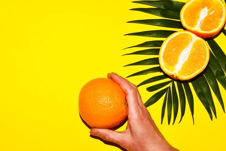 On a tropical leaf lies a cut orange and a hand holds an orange on a yellow background. Place for text. Top view.の写真素材
