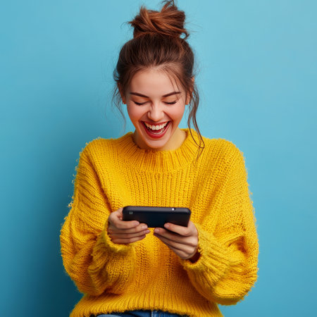 Portrait of happy young woman in yellow sweater using mobile phone on blue backgroundの素材
