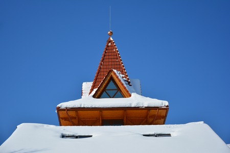 Attic window of triangular shape on snow-covered roof on background of bright blue sky. Sunny winter day.の写真素材