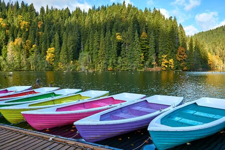 Multi-colored boats at the pier on Lake Lacu Rosu, Romania. Autumn landscape of mountain lake, forest and mountains.の写真素材