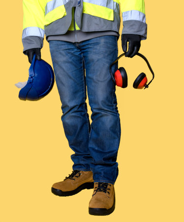 Workman  wearing jeans, gloves, boots and holding protective equipment, helmet and ear defender   on yellow backgroundの写真素材
