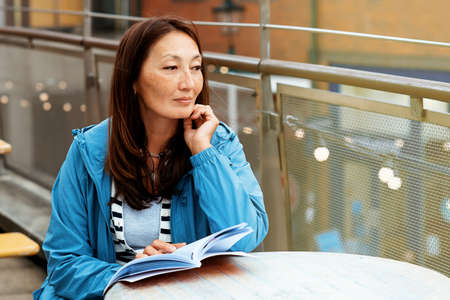 Asian woman in a blue coat sitting in the cafe in the street and reading a bookの写真素材
