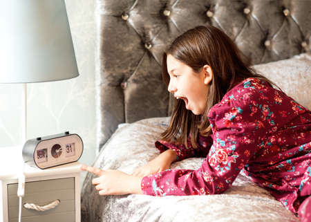 A teenager in pajamas lying in bed and looking at a clock. She is late for school.の写真素材