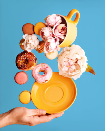 still life of peons in yellow cup, donuts, macaroons, peony and hand holding  saucer flying over a...