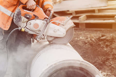 A worker at the construction site cutting a concrete drainage pipe with a petrol concrete saw. Builder covered in a hazardous dust cloud as safety procedure breached on construction site.の写真素材