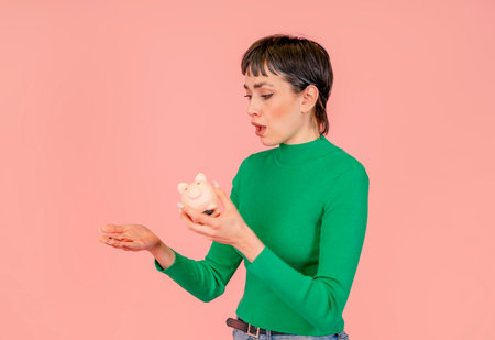 Sad woman wearing green casual sweater discovering empty piggy bank without savings. Investment, saving money, currency, deposit. Indoor studio shot isolated on pink background.の写真素材