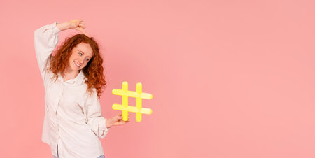 Excited woman in white shirt holding yellow sign hashtag in her hand on pink background with copy space. social media, blogging and viral topics on internet conceptの写真素材