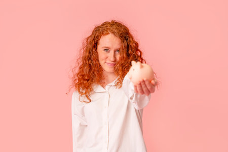 happy red hair woman in white shirt saving money for rainy day in the piggy bank, investment, saving money, currency, deposit. Indoor studio shot isolated with copy space on pink background.の写真素材