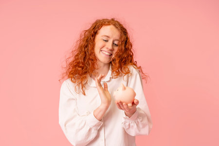 happy red hair woman in white shirt saving money for rainy day in the piggy bank, investment, saving money, currency, deposit. Indoor studio shot isolated with copy space on pink background.の写真素材