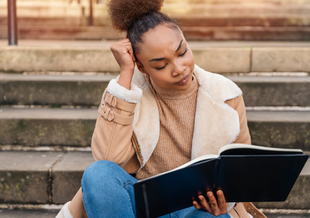 wistful concentrating African Ethnicity woman learning, reading, studying sitting outdoors on stairs in the city. Education conceptの写真素材