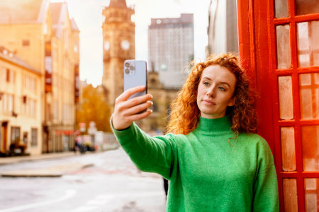 Young woman portrait close to red telephone box in English city taking a selfie with smart phone. Old city in the background.の写真素材