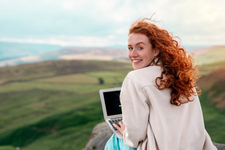 enjoying redhead woman reaching the destination, using laptop, and enjoying mountains landscape on the top of mountain at sunset. Enjoy hiking and exploring new places conceptの写真素材