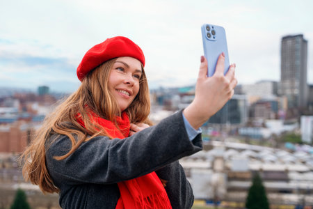 happy woman in a grey coat, red scarf and beret sitting in front of the city Sheffield and taking photos on a great spring dayの写真素材