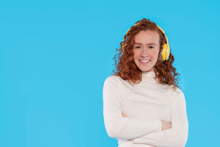 Happy young redhead woman carried away with music dances carefree with arms raised with stereo headphones on her ears dressed in jeans and white shirt on a blue backgroundの写真素材