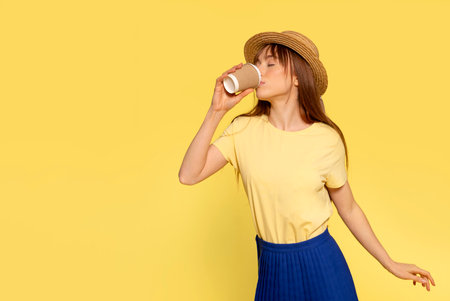 Happy young woman holding and drinking from a cup of coffee or tea on yellow backgroundの写真素材