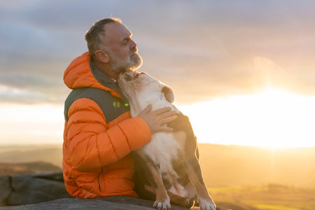 senior mature bearded man with english bulldog sitting on top of mountain at sunsetの写真素材
