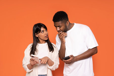 serious woman pointing at screen, man with hand on cheek looking at phone and thinking on orange backgroundの写真素材