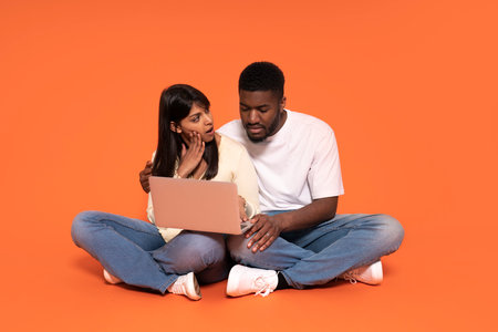 woman cross-legged in shock pointing at screen, man looking at laptop and thinking on orange backgroundの写真素材