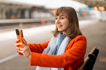 joyful young lady using phone taking selfie waiting for train at railway station on platformの写真素材