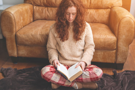 focused redhead young woman studying reading book sitting on floor near sofa at home Toned imageの写真素材