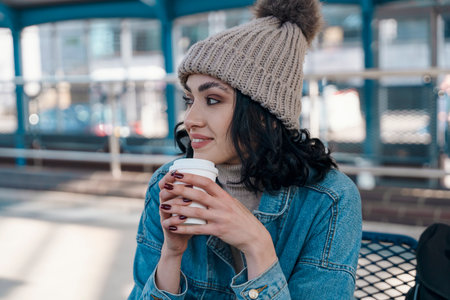 happy young woman drinking take away coffee and waiting for bus at bus stationの写真素材
