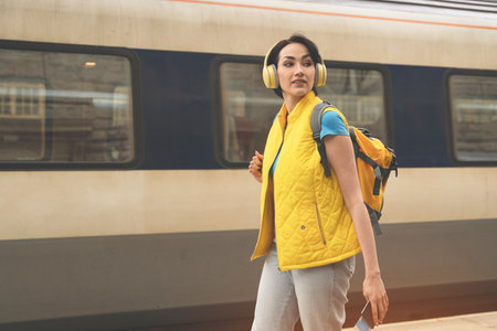 young female traveler, with backpack and headphones passport tickets waiting for train on platform at railway station toned imageの写真素材