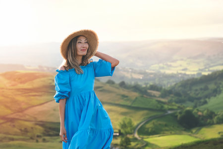joyful Asian woman in dress and hat on top of mountain at summer sunsetの写真素材