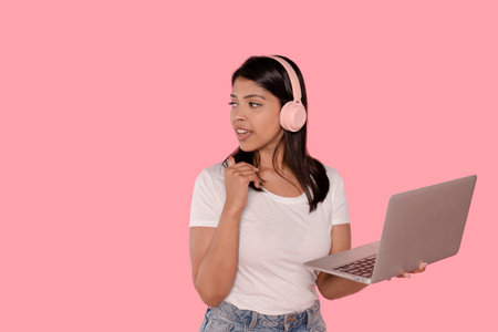 happy Indian woman with headphones using laptop looking away on pink background with copy spaceの写真素材