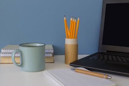 Front view on modern creative workspace. Laptop with black screen, blue cup of coffee, yellow pencils on reusable paper tube on white table. Home office of a creative entrepreneur or studentの写真素材