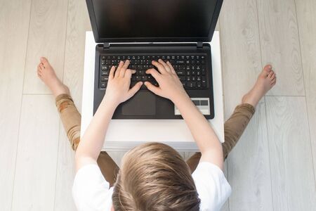 Top view on child boy sitting on white wooden floor typing on his laptop, studying online at home or having rest. Unusual composition with copy spaceの写真素材