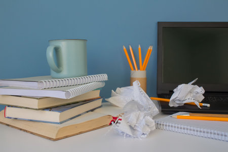 Front view on chaos or mess on white table with black laptop, books, cup of coffee and yellow...