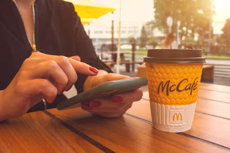 Lviv, Ukraine - June 14, 2020 : young woman with perfect manicure scrolling feed with fresh news or social media and drinking coffee during breakfast or lunch at McDonald's restaurant. Early morningのeditorial素材