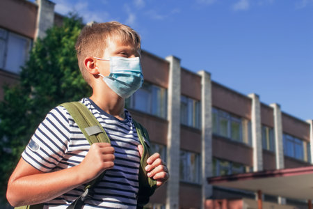 Boy wearing protective disposable face mask, standing in front of school building. Back to school after quarantine and pandemic prevention conceptの写真素材