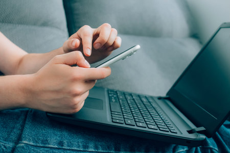Closeup view of young woman hands working remotely from home, using both laptop and smartphone....