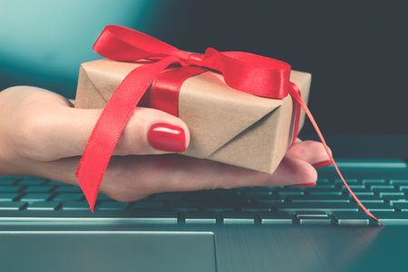 Womans hand holding present box with red bow and laptoptop computer as background behind it for your...