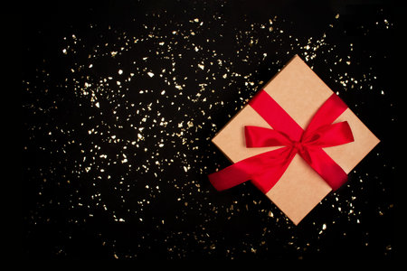 Present box with red bow on luxury black background with golden sparkles around and copyspace for...