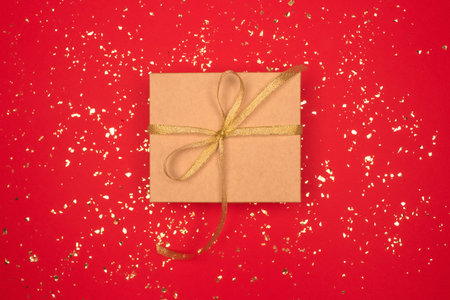 Gift box with golden tape bow on festive red background with golden sparkles around and copyspace...