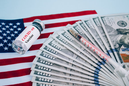 Dollars bills and vial dose of COVID-19 vaccine with syringe against USA flag and blue background with copy space - global vaccination and money abuse concept. Covid vaccine cost. Selective focusの写真素材